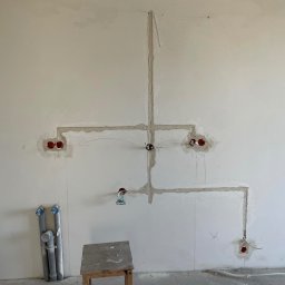 Dominik Narloch - Ściana w trakcie prac elektrycznych: wyfrezowane bruzdy zalane gipsem, widoczne puszki instalacyjne i rury PCV przy podłodze, krzesło robocze w rogu pomieszczenia.