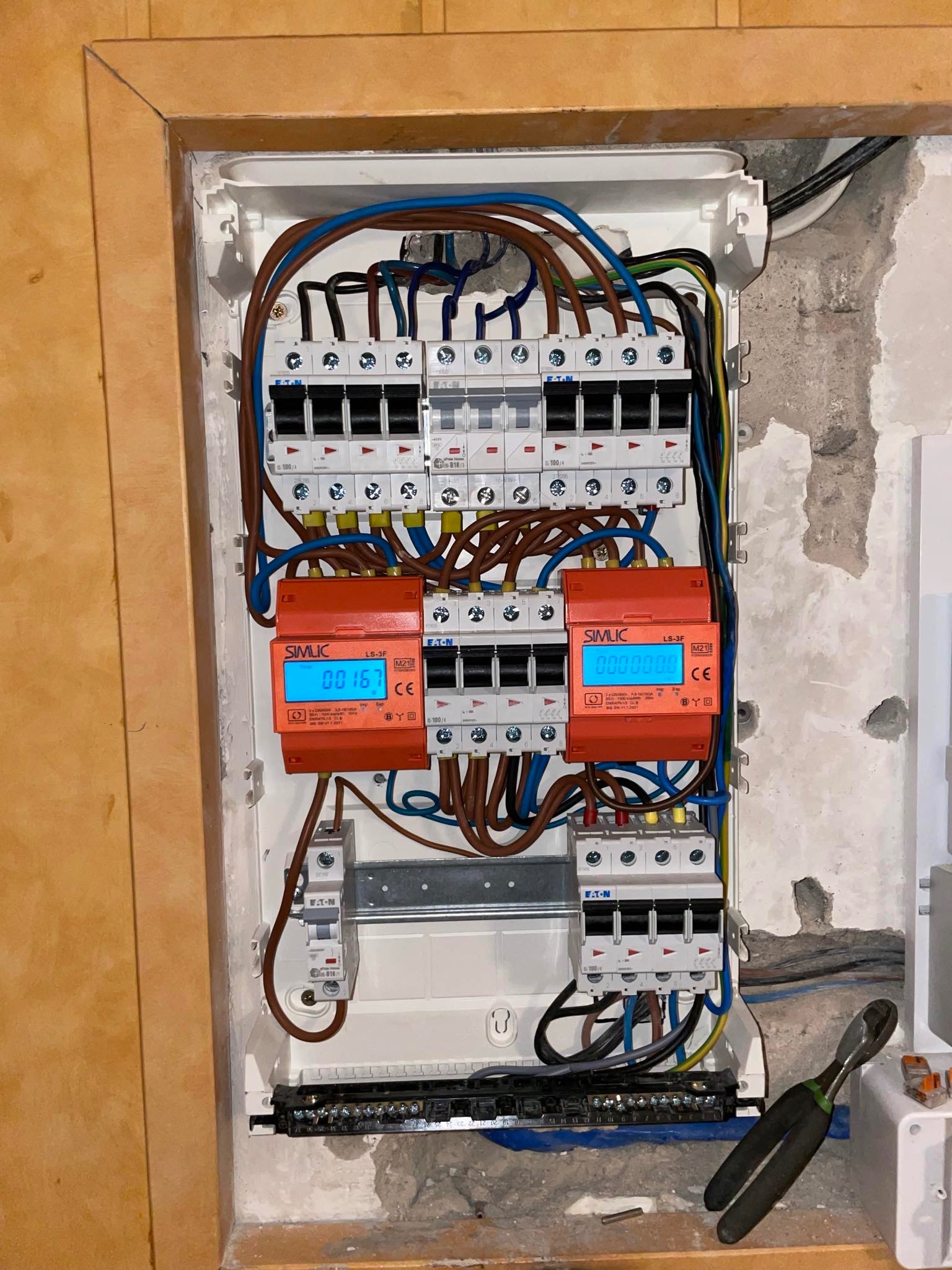 Otwarta skrzynka rozdzielcza z bezpiecznikami firmy Eaton i pomarańczowymi licznikami zużycia energii Simlic z wyświetlaczami LCD, widoczne przewody w różnych kolorach oraz narzędzia elektryczne obok.