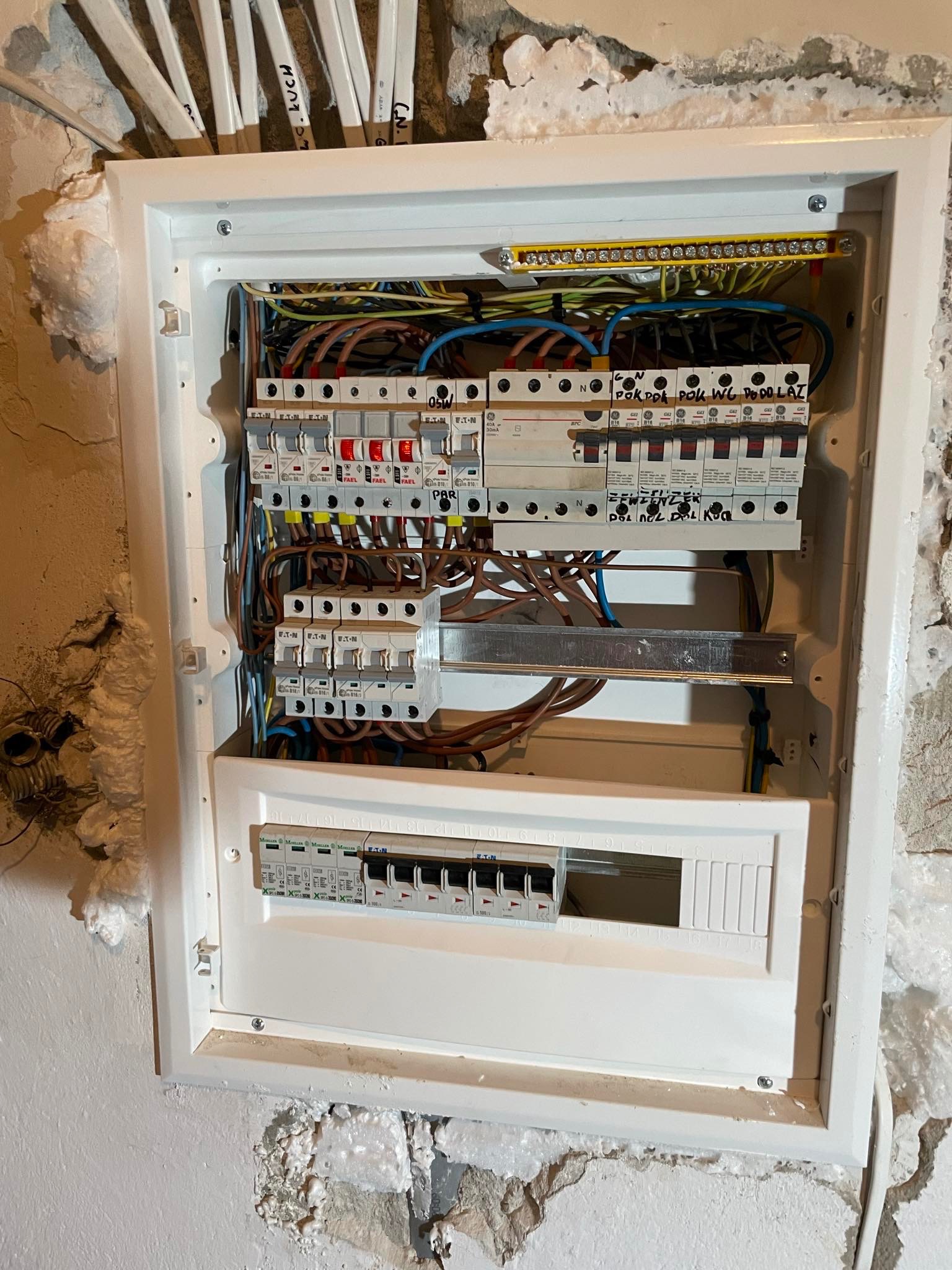 Otwarta skrzynka bezpiecznikowa z widocznymi przewodami, bezpiecznikami i listwami zaciskowymi, zamontowana na nierównej ścianie z resztkami zaprawy i pianki montażowej.