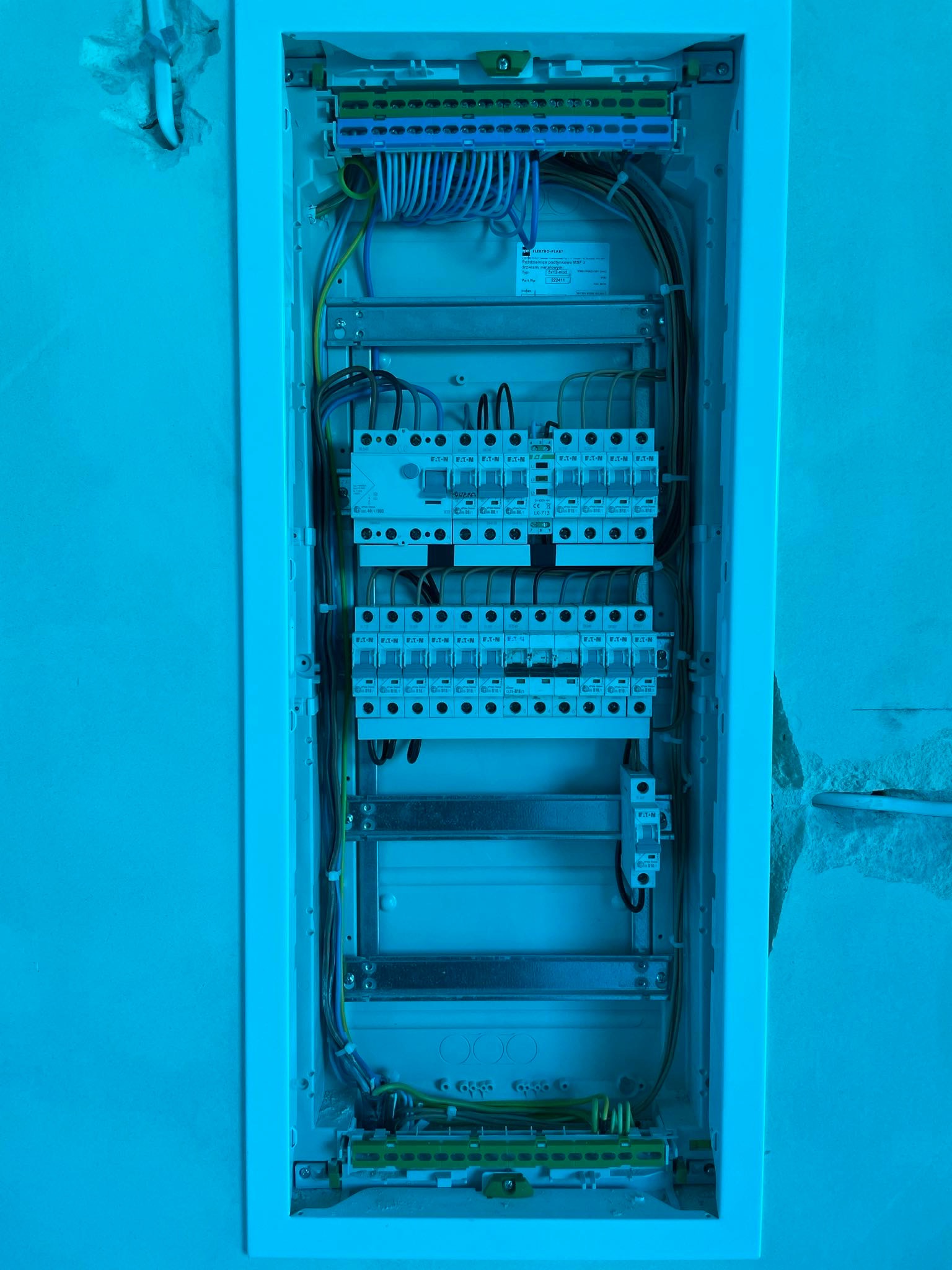 Wewnętrzny widok skrzynki rozdzielczej podtynkowej z zainstalowanymi bezpiecznikami EATON, przewodami i listwami zaciskowymi, widoczne kable wychodzące ze ściany.