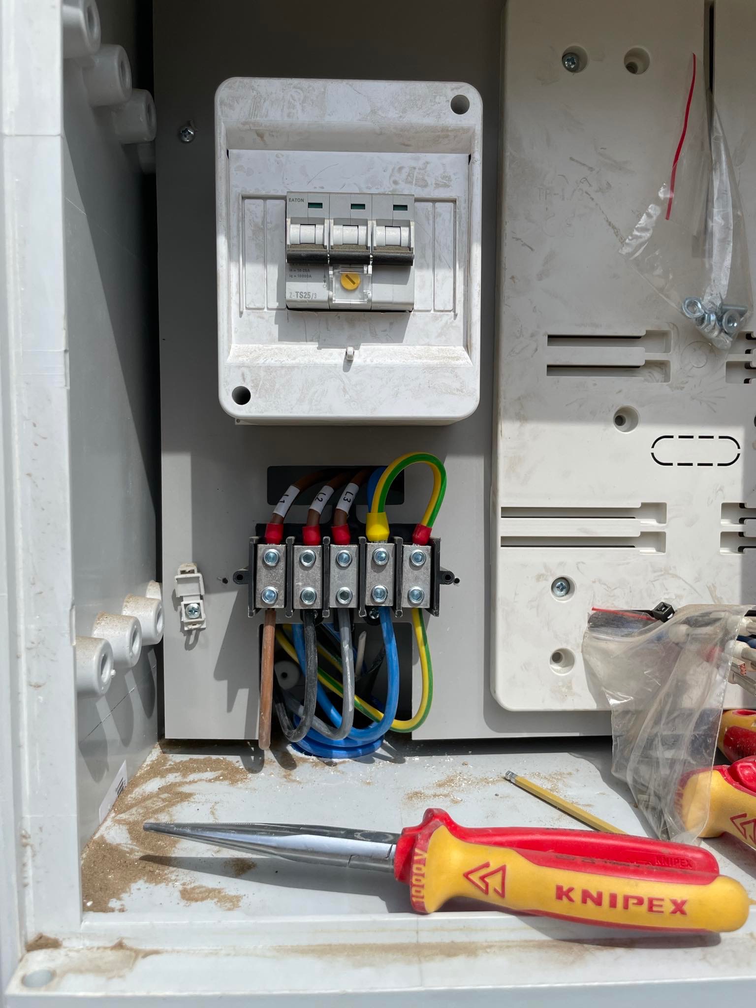Otwarta skrzynka elektryczna z bezpiecznikami, złączkami kablowymi i narzędziem Knipex, widoczne okablowanie z oznaczeniami L1, L2, L3, oraz przewód ochronny żółto-zielony, całość lekko zabrudzona.