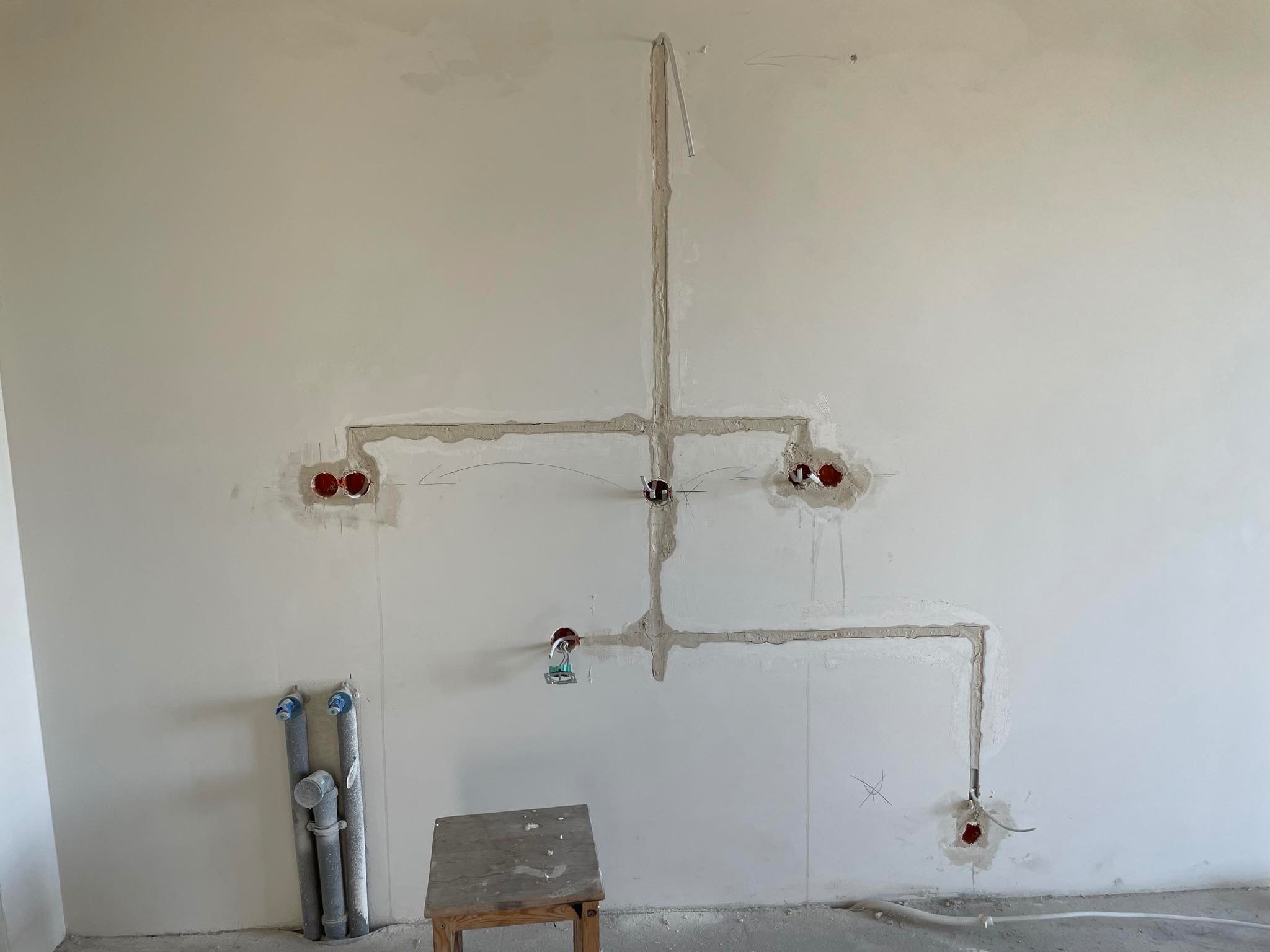 Ściana w trakcie prac elektrycznych: wyfrezowane bruzdy zalane gipsem, widoczne puszki instalacyjne i rury PCV przy podłodze, krzesło robocze w rogu pomieszczenia.
