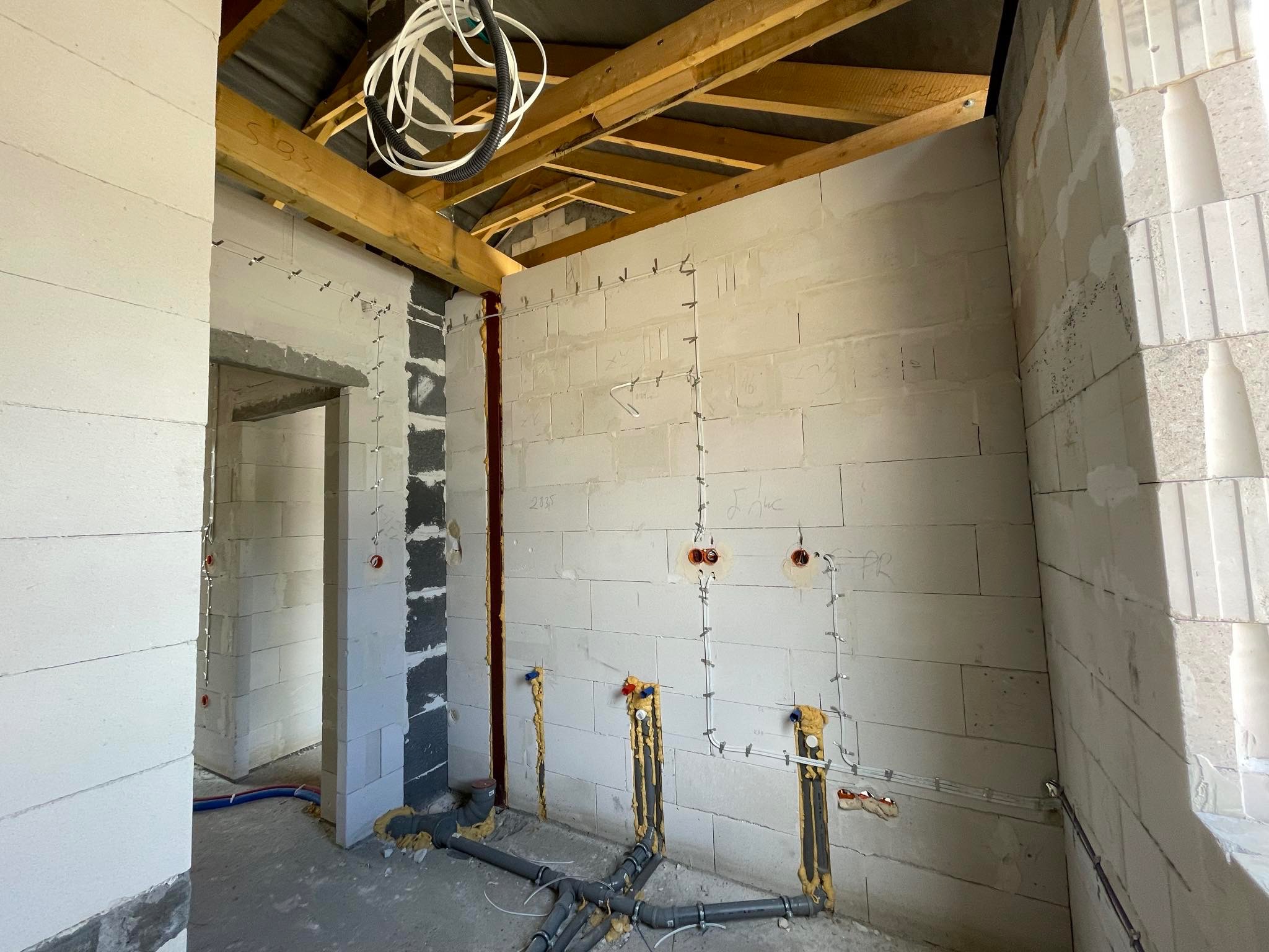Wnętrze w stanie surowym, ściany z białych bloczków, widoczne rozprowadzone przewody elektryczne mocowane do ściany klamrami, instalacja wodno-kanalizacyjna w podłodze, drewniana konstrukcja dachu...
