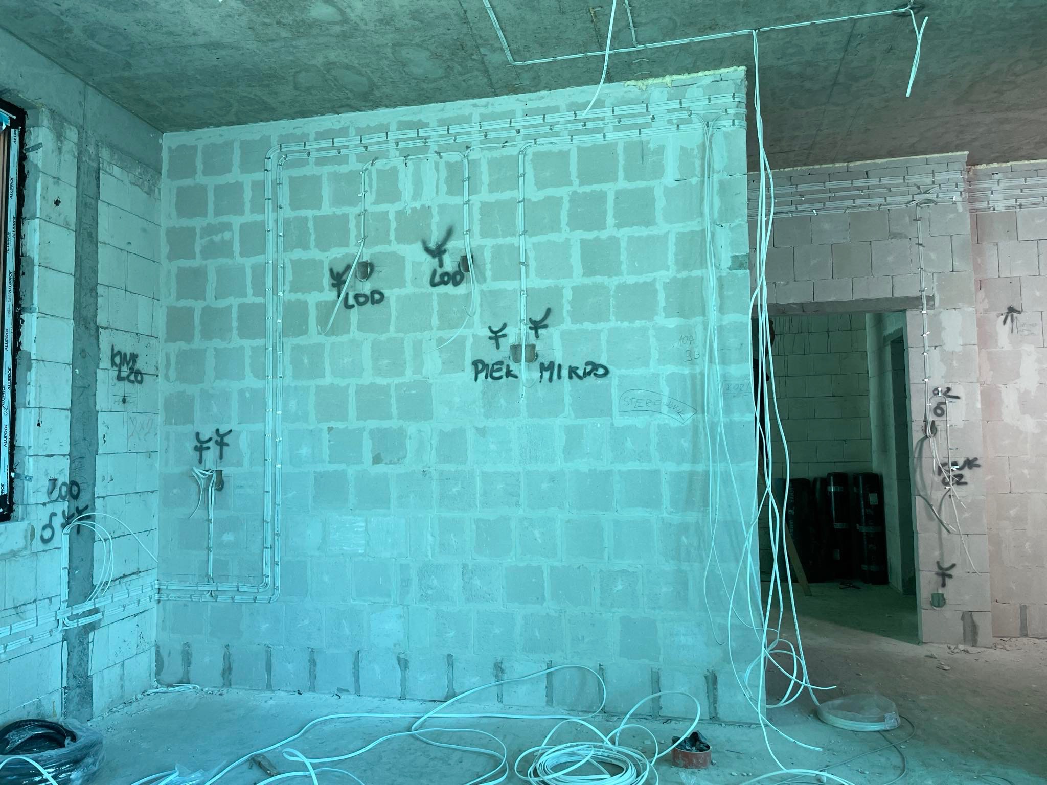 Surowy stan wnętrza z widocznymi przewodami elektrycznymi w listwach na ścianach z betonu komórkowego, napisy wykonane sprayem na ścianie, instalacja elektryczna w trakcie montażu.