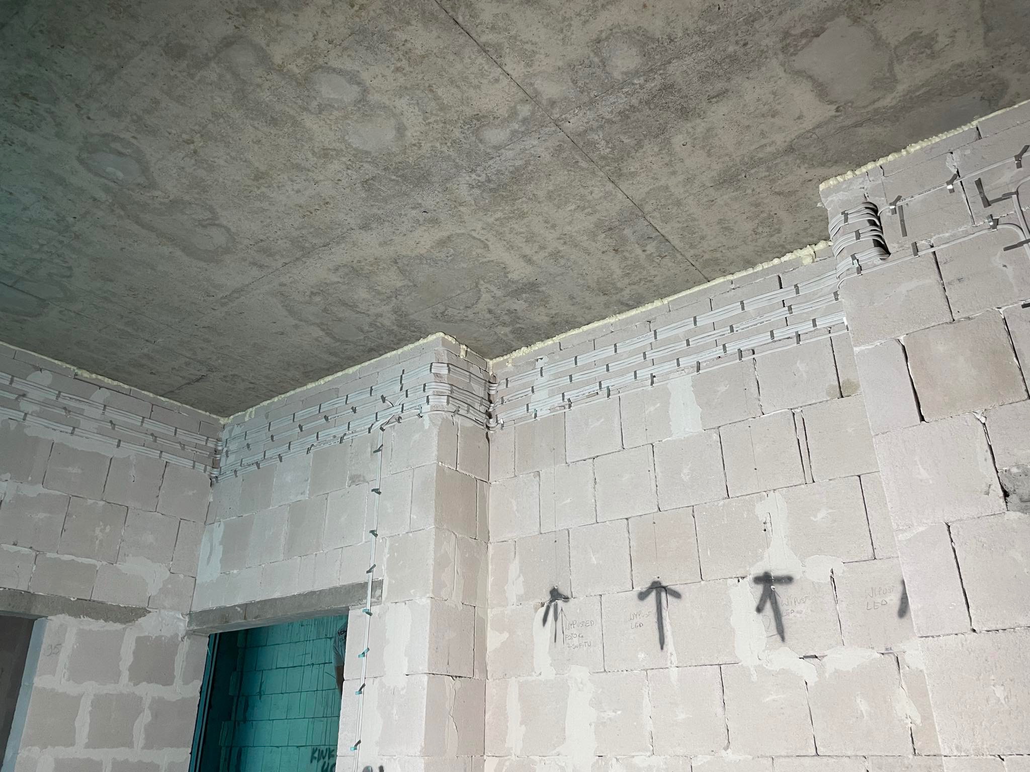 Surowy stan wnętrza w trakcie prac: widoczne kable w korytkach przymocowane do ściany z bloczków, surowy betonowy sufit, pianka montażowa w szczelinach, strzałki z opisami na ścianie.
