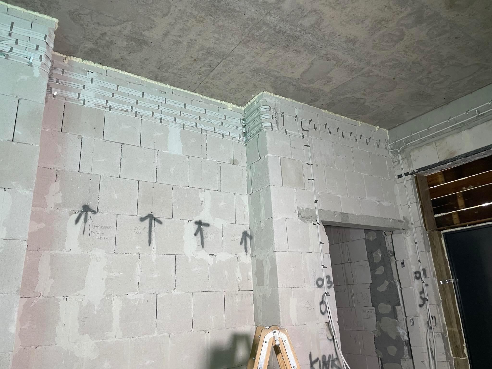 Surowy stan: ściana z betonu komórkowego z rozprowadzonymi przewodami elektrycznymi, zabezpieczonymi pianką montażową pod sufitem, oznaczenia na ścianie, widoczna drabina.