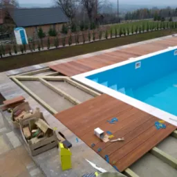 Montaż drewnianego tarasu wokół basenu, widoczne deski, konstrukcja nośna, poziomica, pędzle, pudełka po akcesoriach i narzędziach, w tle ogród i zabudowania.