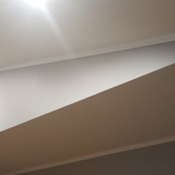 MarHor Renovations - Wykończone wnętrze z podwieszanym sufitem i oświetleniem punktowym, widok na fragment ściany pomalowanej na szaro i część drzwi z dekoracyjną ramą.