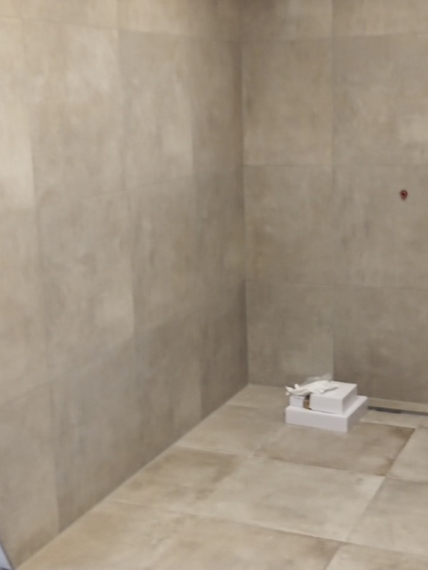 Wykończona kabina prysznicowa z beżowymi płytkami, widoczne pudełka po armaturze na podłodze, ślad po wierceniu w ścianie.