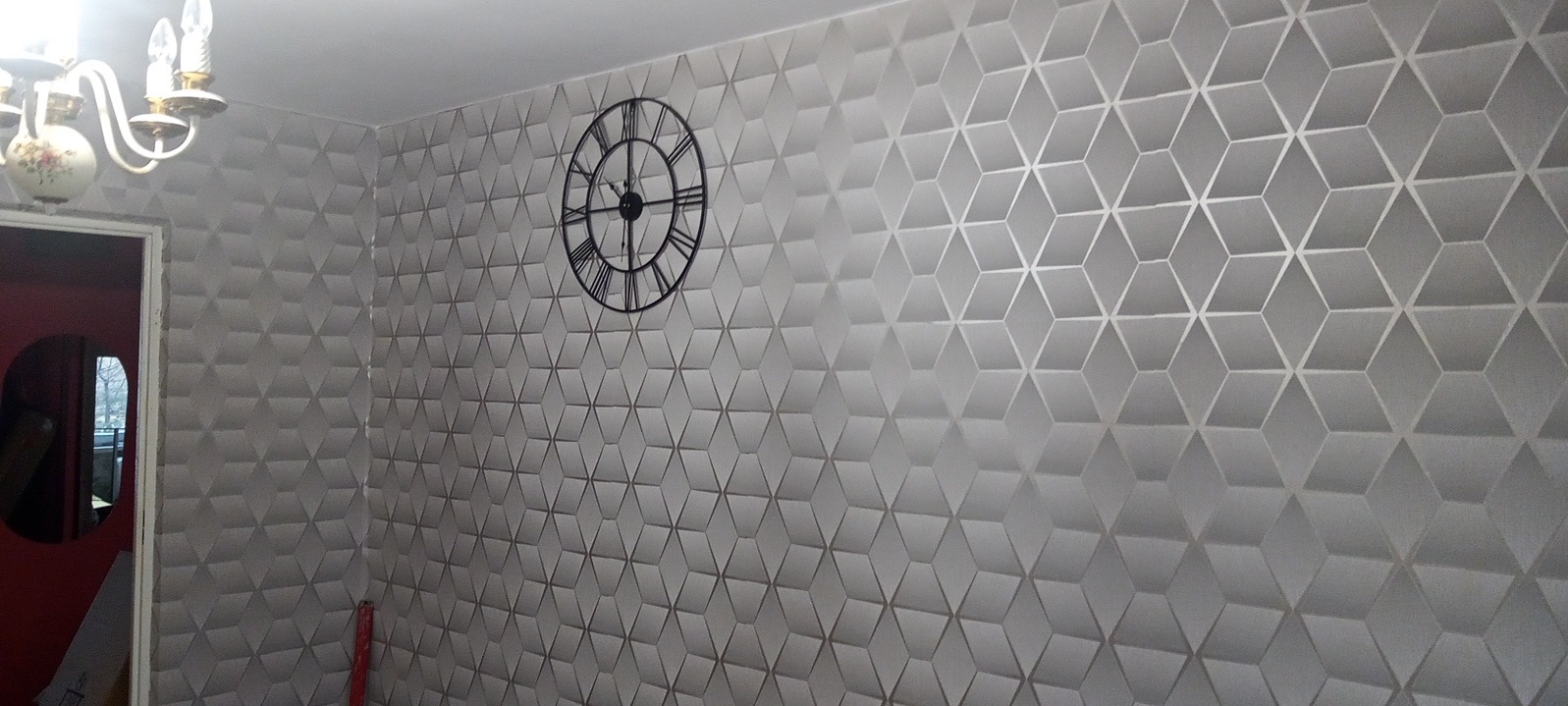 Szara ściana w pokoju z trójwymiarową tapetą w geometryczne wzory, ozdobiona dużym, okrągłym zegarem wiszącym i oświetlona przez żyrandol z kwiatowym motywem, widoczny fragment czerwonego pokoju obok.