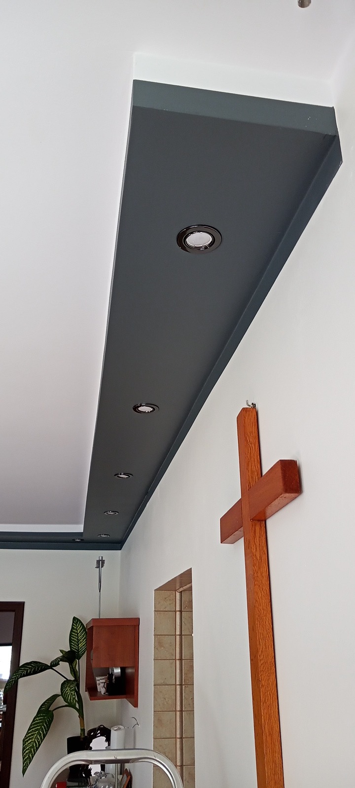 Pomalowany na szaro fragment sufitu z zamontowanymi punktowymi lampami LED, widoczny drewniany krzyż na ścianie i fragment drabiny malarskiej.