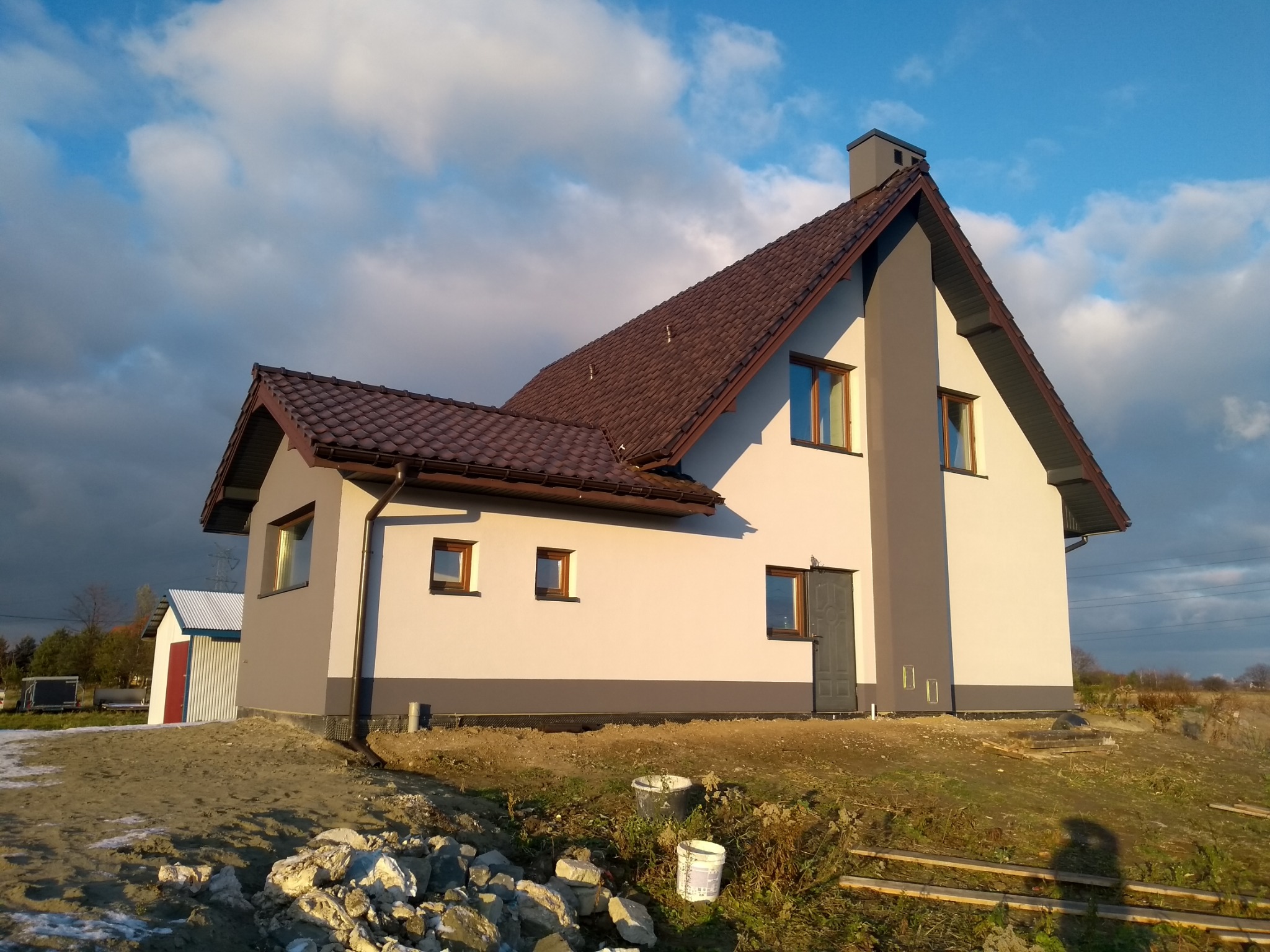 Dwukondygnacyjny dom jednorodzinny z brązową dachówką i szarymi akcentami elewacji, widoczny w słoneczny dzień na tle lekko zachmurzonego nieba, z widocznymi elementami przygotowania terenu wokół.