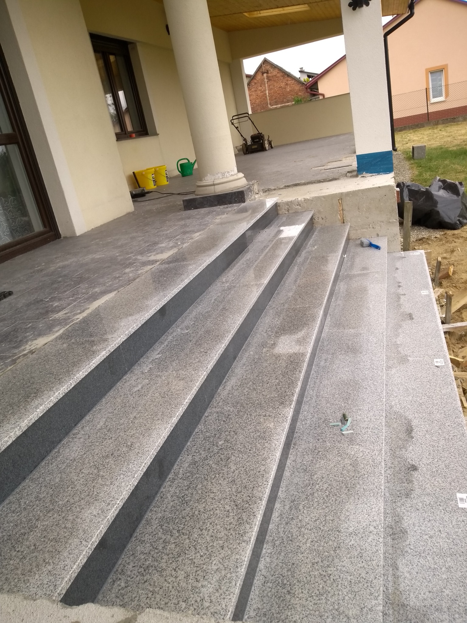 Granitowe schody zewnętrzne z widocznymi śladami prac montażowych, w tle kolumny werandy i trawnik z kosiarką.