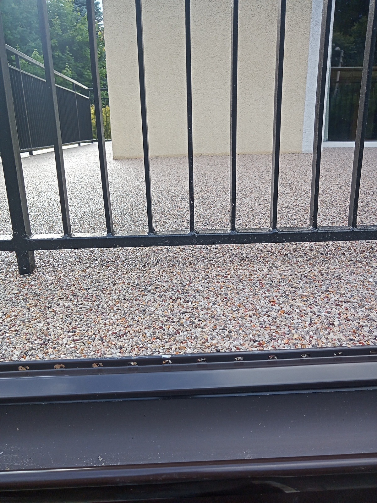 Balkon z żywicy kamiennej widziany przez czarne metalowe barierki, z widoczną fakturą posadzki i fragmentem elewacji budynku w tle.