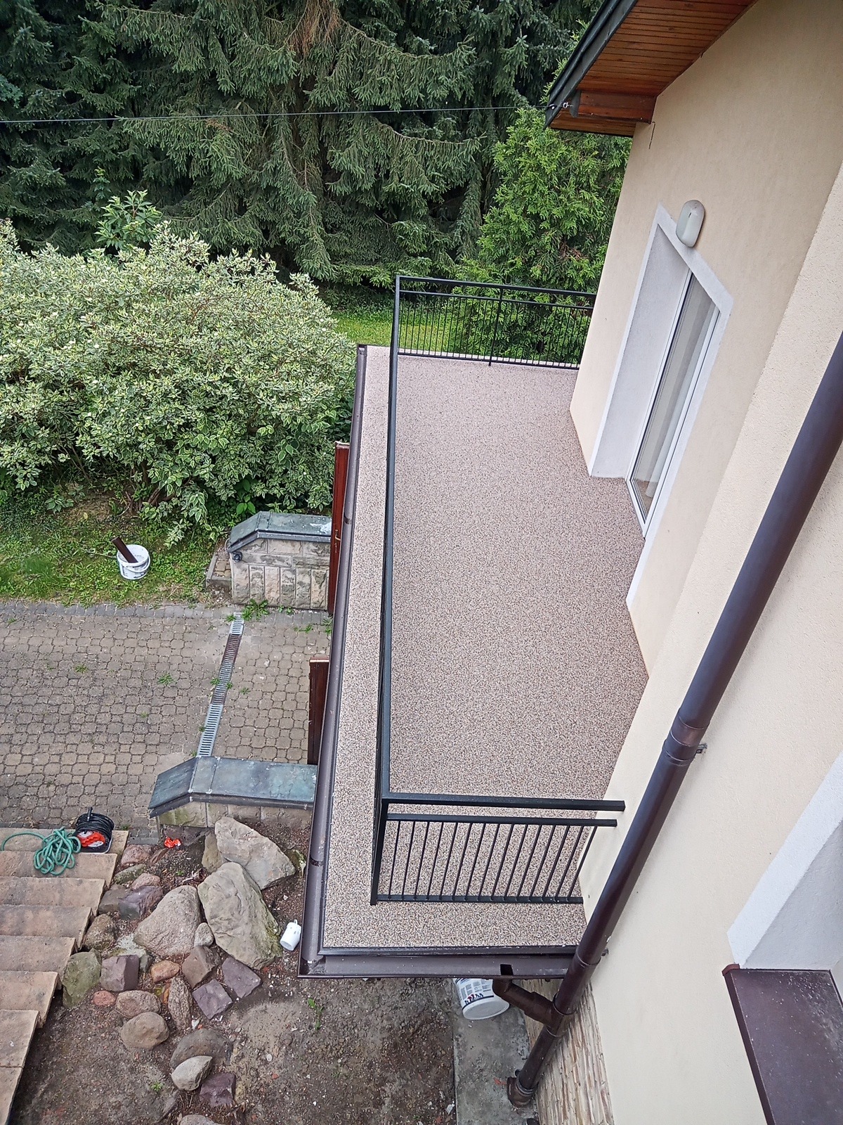 Widok z góry na balkon z nową, beżową, żywiczną posadzką, czarną balustradą i brązową rynną, z widocznymi elementami otoczenia budynku.