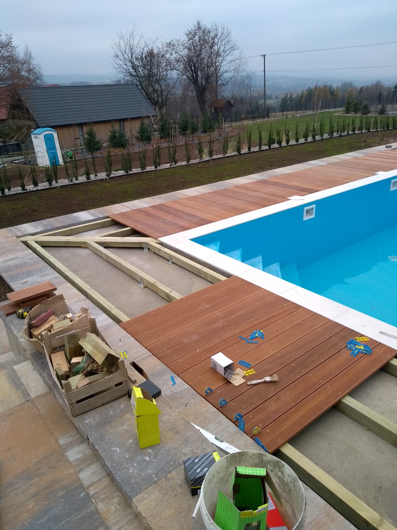 Montaż drewnianego tarasu wokół basenu, widoczne deski, konstrukcja nośna, poziomica, pędzle, pudełka po akcesoriach i narzędziach, w tle ogród i zabudowania.