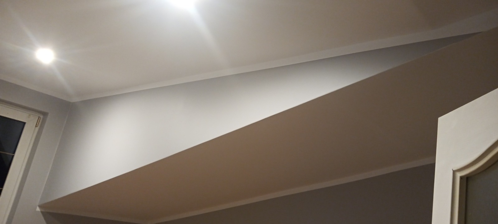 Wykończone wnętrze z podwieszanym sufitem i oświetleniem punktowym, widok na fragment ściany pomalowanej na szaro i część drzwi z dekoracyjną ramą.