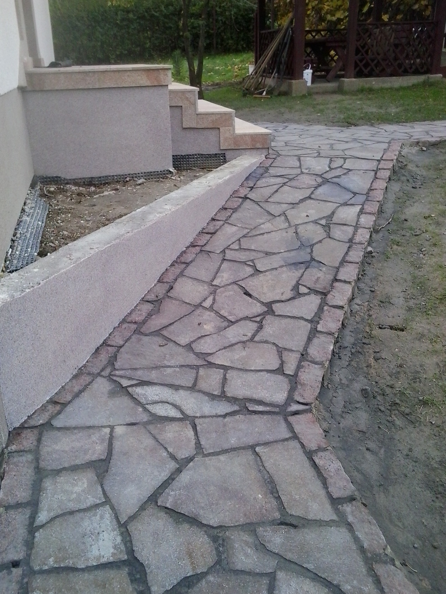 Ułożona ścieżka z nieregularnych kamieni w odcieniach szarości, obrzeżona ceglaną kostką, prowadząca od schodów domu w kierunku altany ogrodowej.