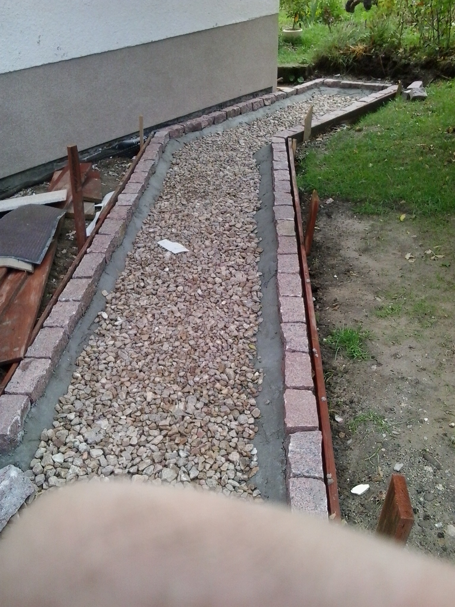 Ścieżka z kamienia otoczona obrzeżem z kostki, w trakcie budowy, widoczne drewniane podpory i zaprawa cementowa, otoczenie z trawnikiem i elementami roślinności.