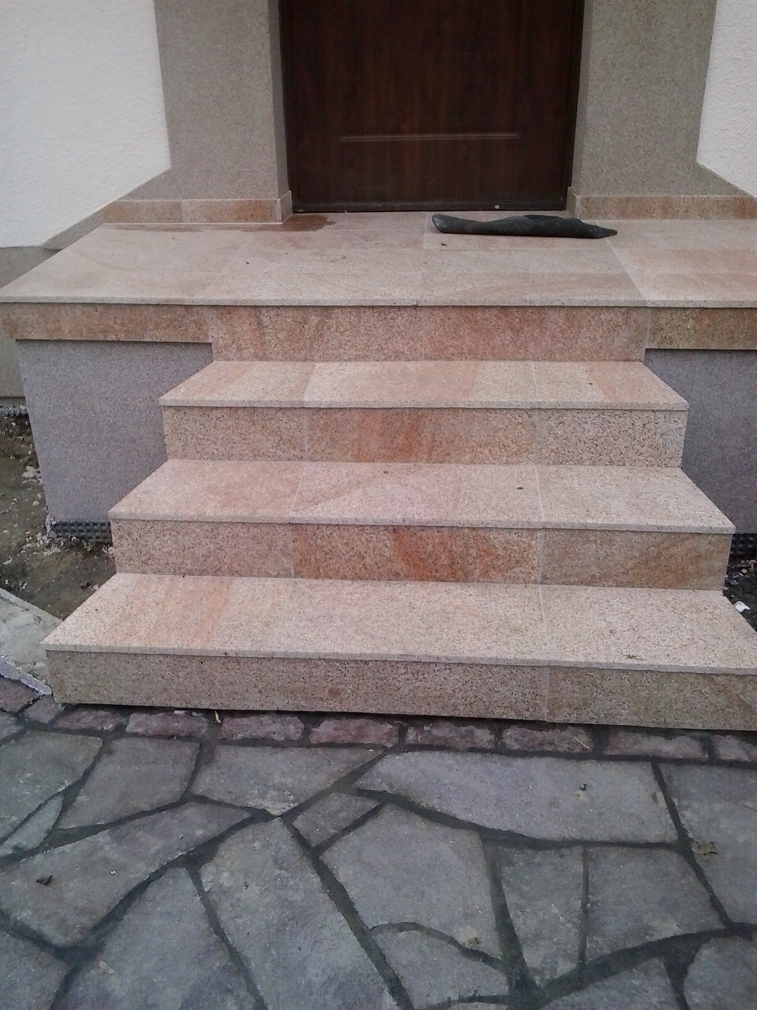 Wejście do domu z czterema schodami wyłożonymi płytkami imitującymi piaskowiec, z ciemnobrązowymi drzwiami i wycieraczką leżącą na górnym podeście.