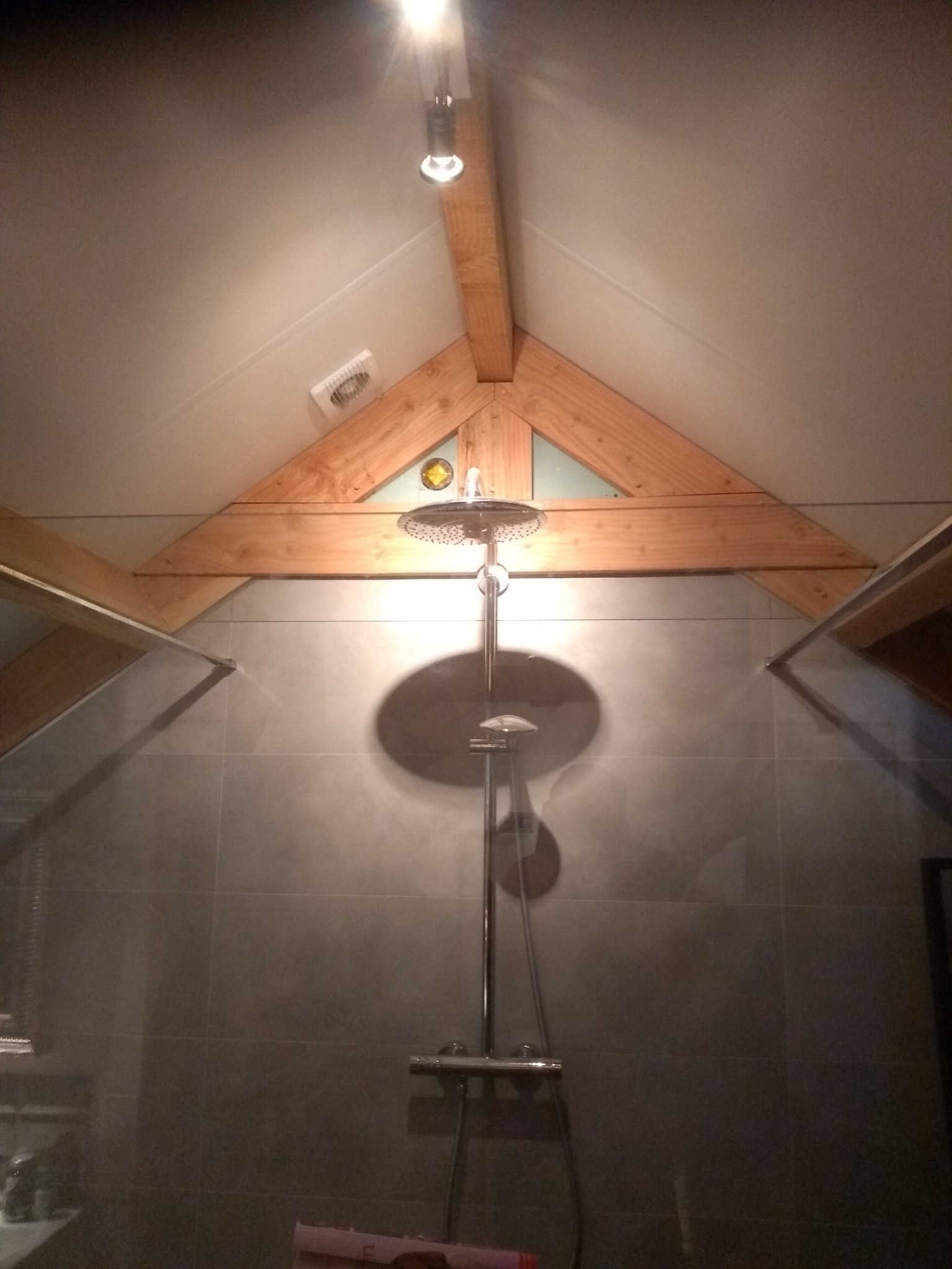Kabina prysznicowa z deszczownicą w łazience na poddaszu, widoczne drewniane elementy konstrukcyjne dachu i szara płytka ceramiczna.