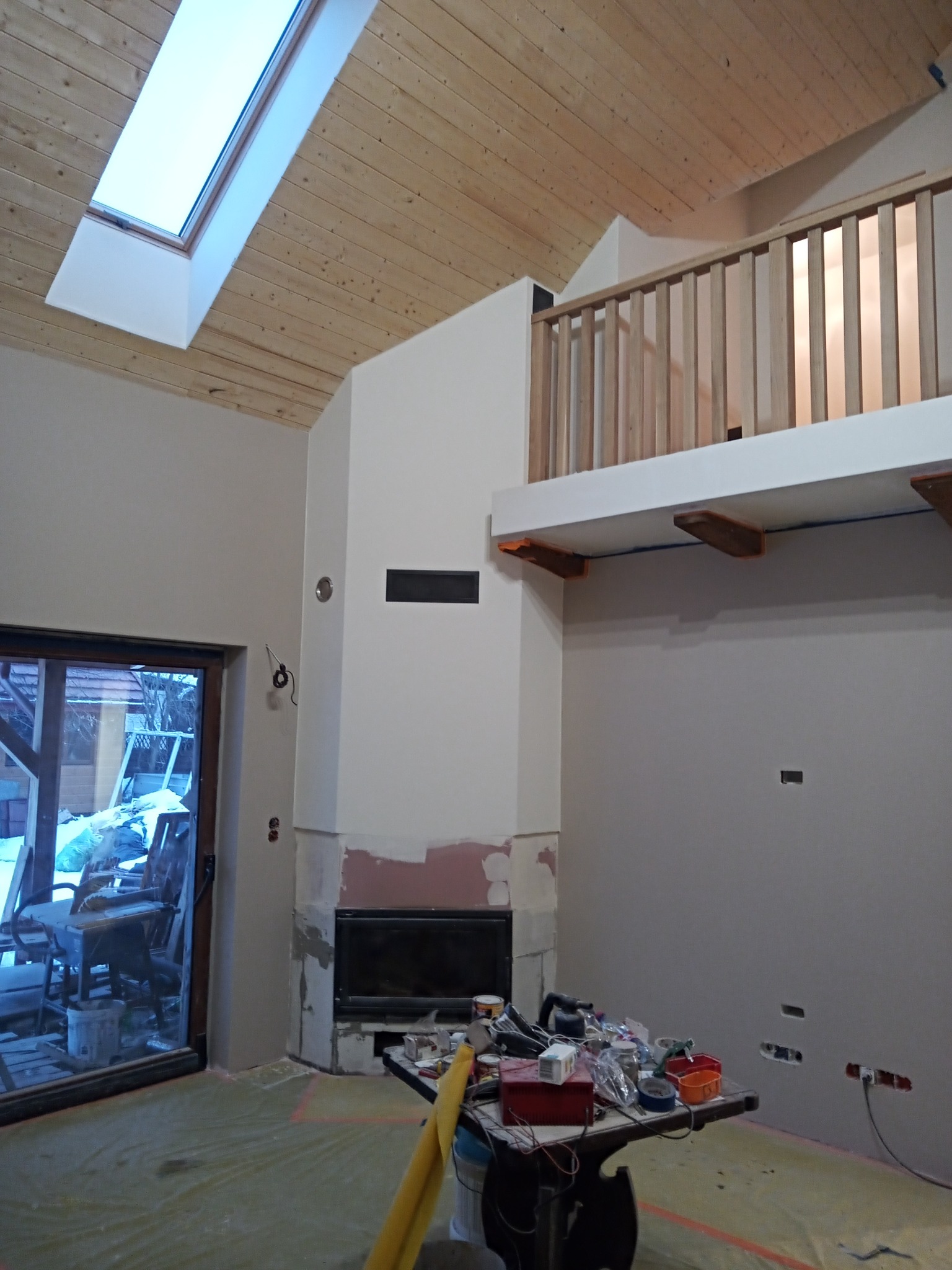 Wnętrze domu w trakcie prac wykończeniowych, widoczny kominek z obudową, drewniany sufit z oknem dachowym, antresola z drewnianą balustradą oraz stół z narzędziami i materiałami budowlanymi.