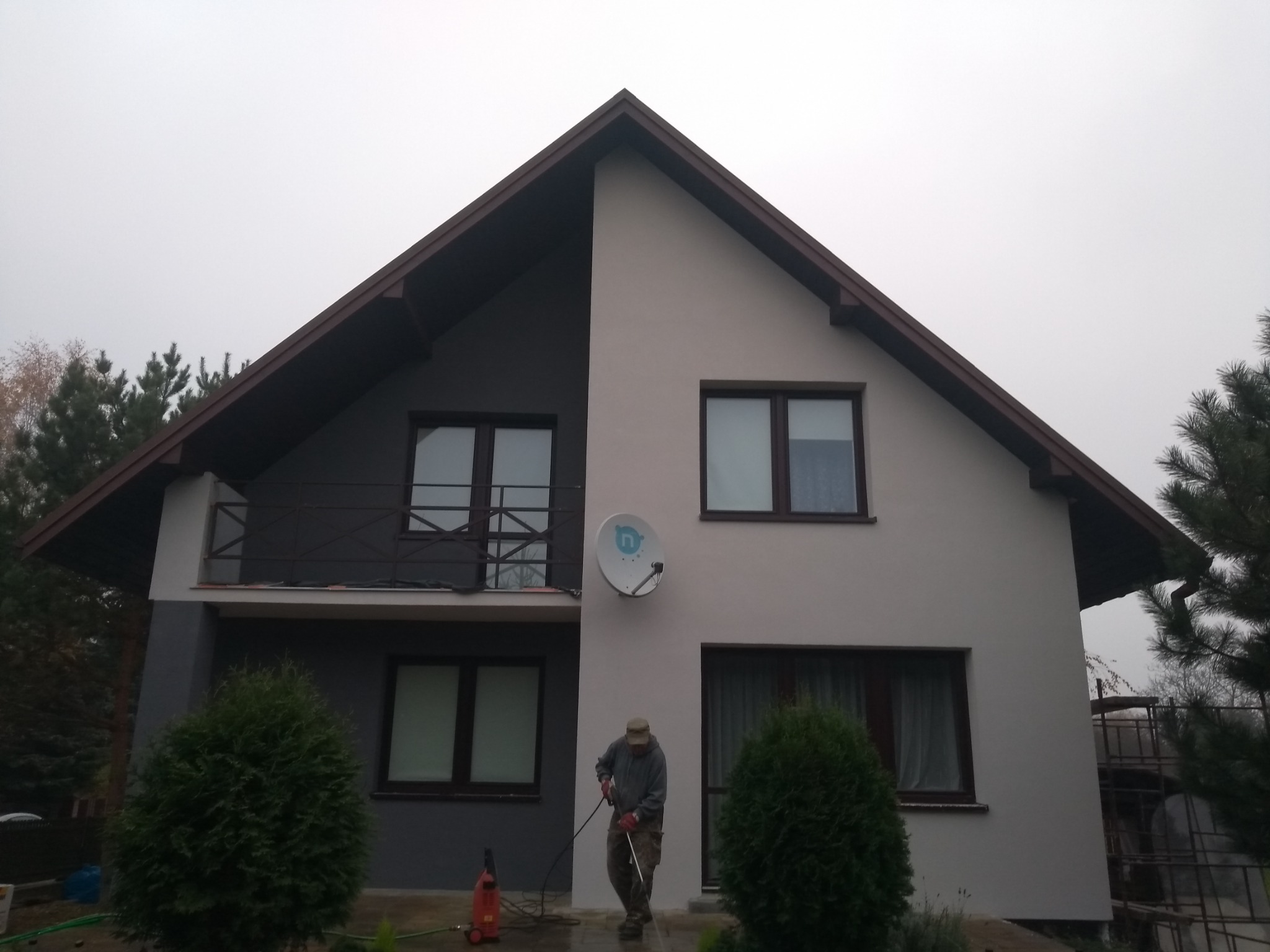 Dwukondygnacyjny dom jednorodzinny z balkonem, czyszczony myjką ciśnieniową przez pracownika w odzieży roboczej, widoczna antena satelitarna na elewacji.