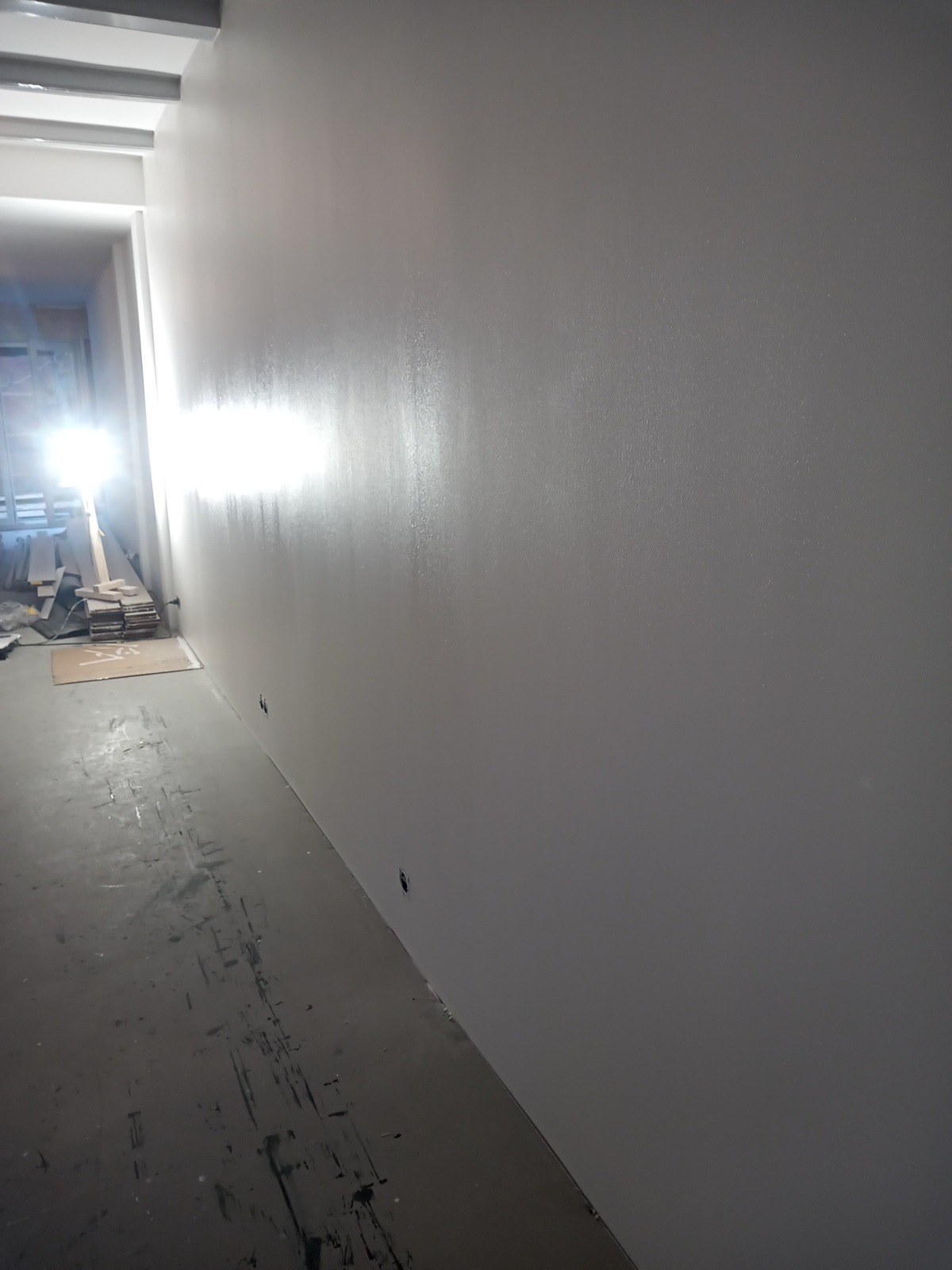 Świeżo pomalowana szara ściana w trakcie prac wykończeniowych, widoczne ślady farby na podłodze i przygotowane deski w oddali, oświetlenie z przodu kadru tworzy blask na ścianie.