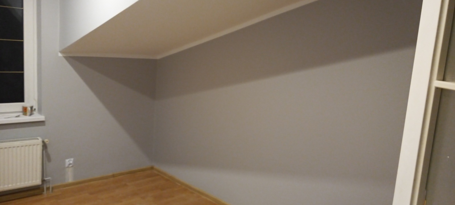 Pomieszczenie po remoncie: ściany pomalowane na szaro, drewniana podłoga, biały grzejnik pod oknem, widoczny skos sufitu.