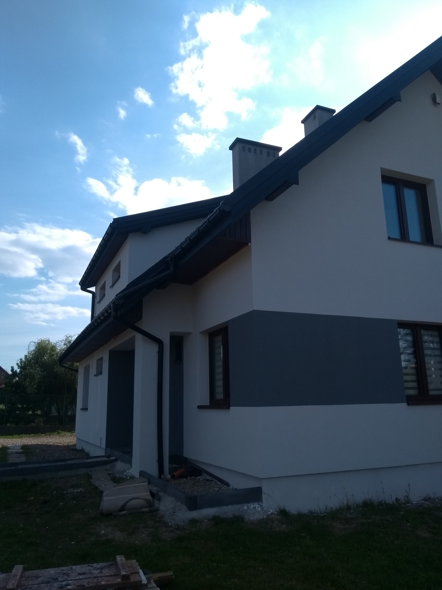 Nowoczesny dom jednorodzinny z dwukolorową elewacją (biało-szarą), ciemną stolarką okienną i dwiema wystającymi częściami kominowymi, sfotografowany w słoneczny dzień z perspektywy lekko od dołu.
