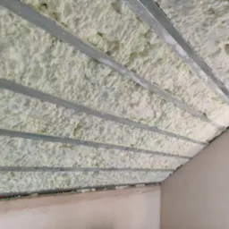 Poddasze ocieplone pianką, widoczne metalowe profile konstrukcyjne, nierównomierna struktura piany, ściana w tle przygotowana do dalszych prac wykończeniowych.