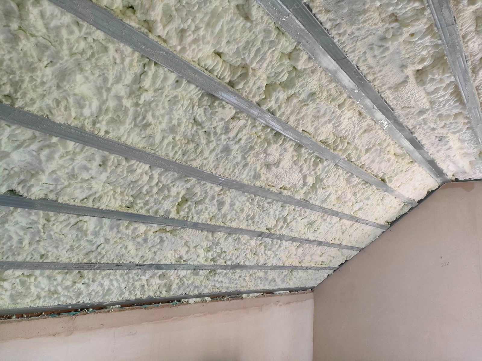 Poddasze ocieplone pianką, widoczne metalowe profile konstrukcyjne, nierównomierna struktura piany, ściana w tle przygotowana do dalszych prac wykończeniowych.