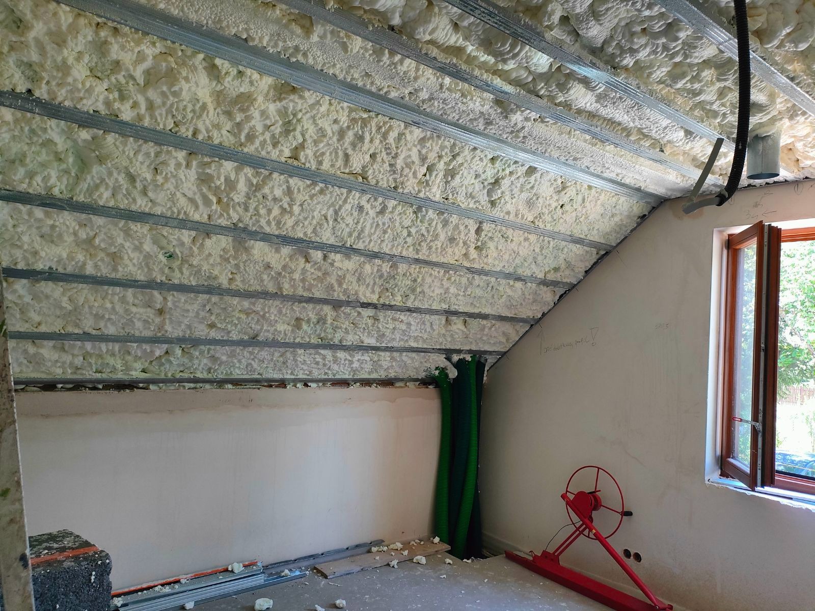 Poddasze ocieplone pianką PUR z widocznymi metalowymi profilami montażowymi, z fragmentem ściany z otwartym oknem i zielonymi rurami wentylacyjnymi w rogu.