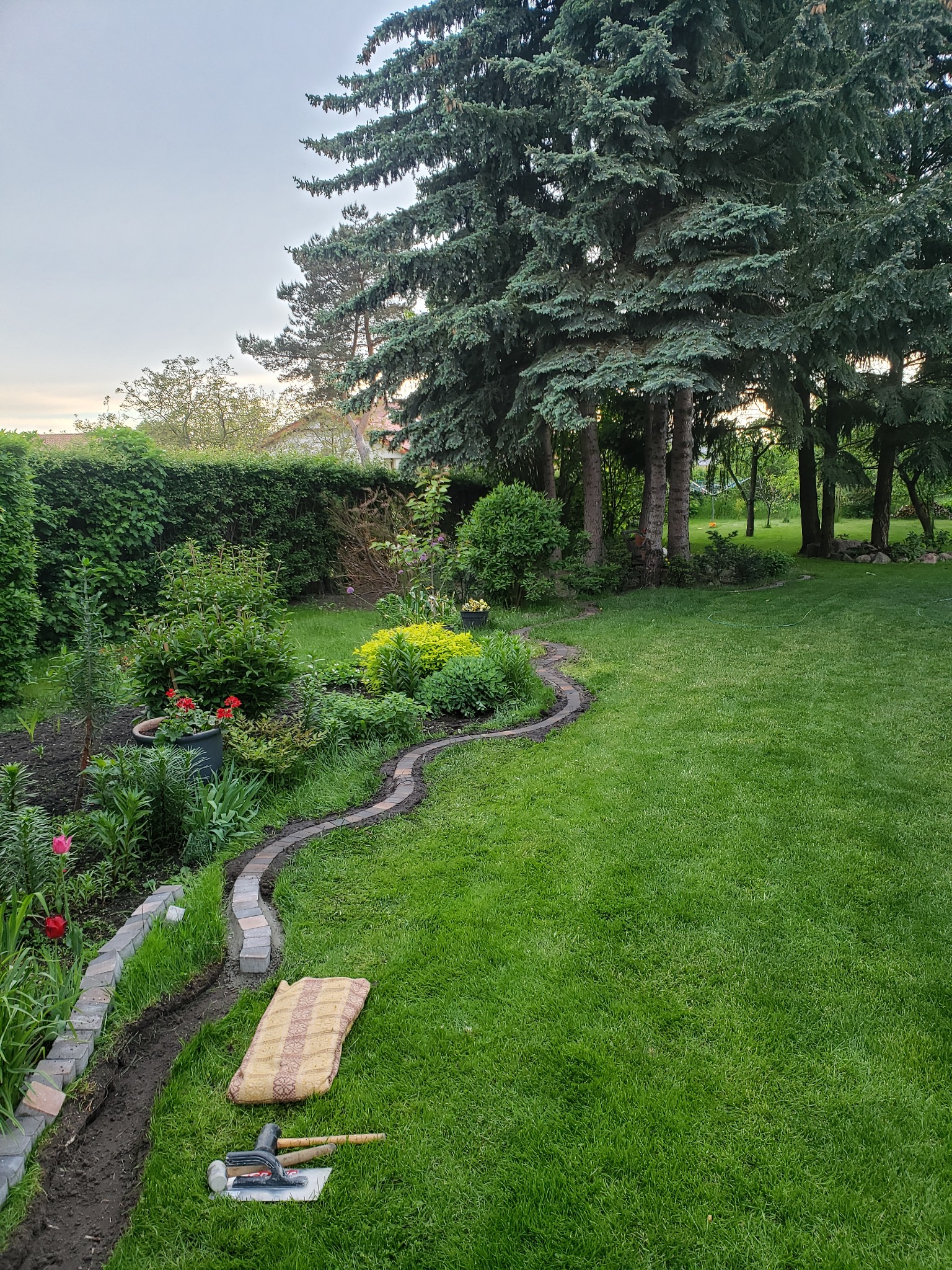 Układanie obrzeża trawnikowego z kostki brukowej w ogrodzie z dużymi drzewami iglastymi w tle; widoczne narzędzia i mata ochronna.