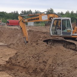 ZECO POZYTYWNA ENERGIA SPÓŁKA Z OGRANICZONĄ ODPOWIEDZIALNOŚCIĄ - Żółta koparka Liebherr 924 w trakcie prac ziemnych, widoczny wykop z betonowymi elementami oraz inne maszyny w tle, dzień pochmurny.