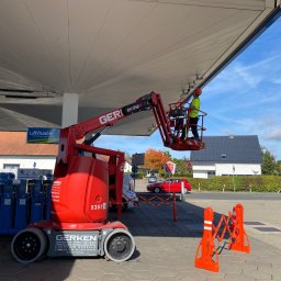ZECO POZYTYWNA ENERGIA SPÓŁKA Z OGRANICZONĄ ODPOWIEDZIALNOŚCIĄ - Pracownik w pomarańczowym kasku i żółtej kamizelce na czerwonym podnośniku koszowym Gerken, wykonujący prace konserwacyjne lub instalacyjne pod zadaszeniem stacji benzynowej Aral w słoneczny dzień.