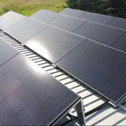 Rząd paneli słonecznych na dachu z blachy trapezowej, widoczne mocowania i kable, w tle zieleń drzew i trawy.