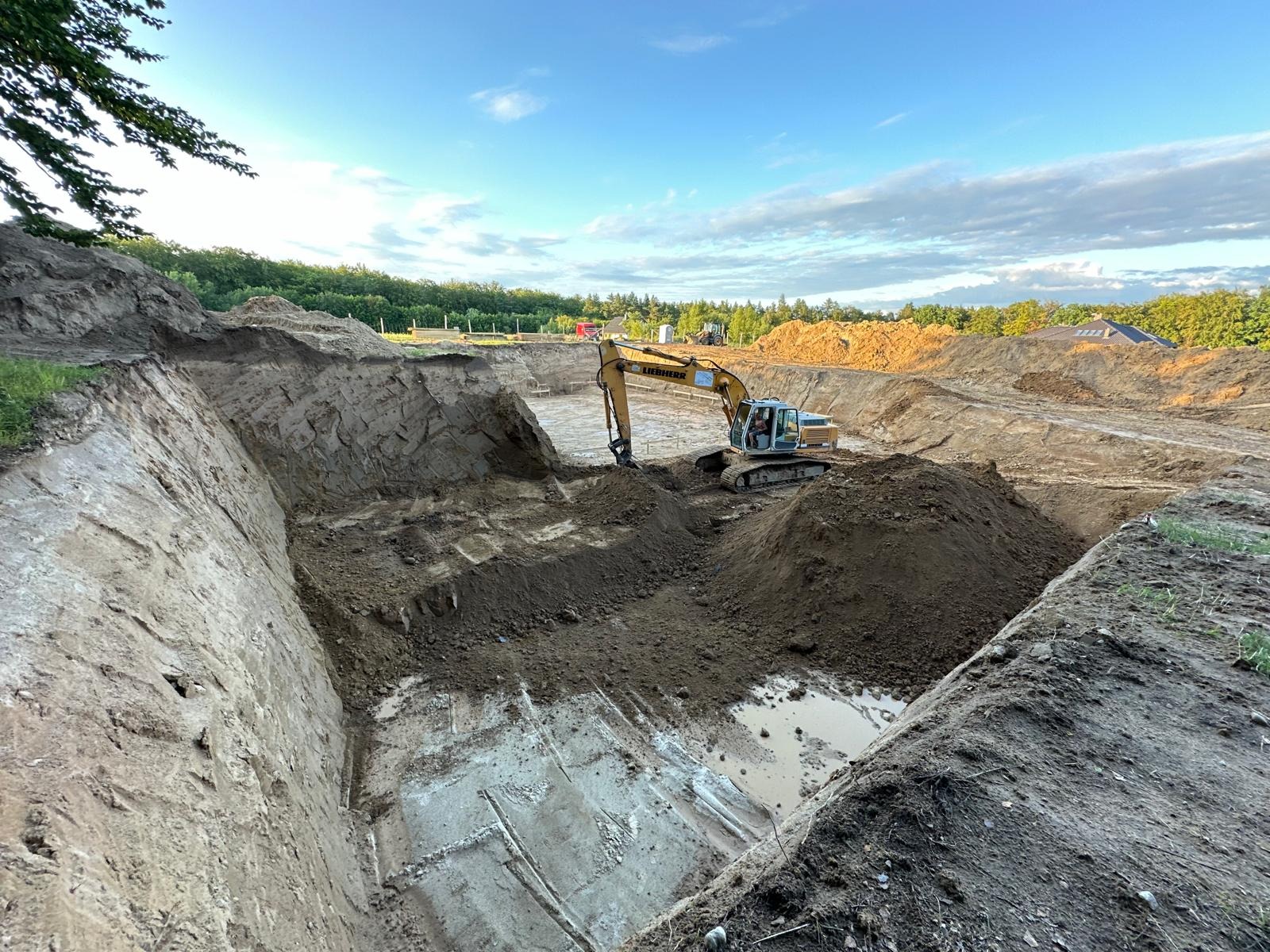 Żółta koparka Liebherr w trakcie prac ziemnych przy wykopie, widoczne ślady po kopaniu na dnie wykopu z kałużami wody, w tle drzewa i błękitne niebo z chmurami.