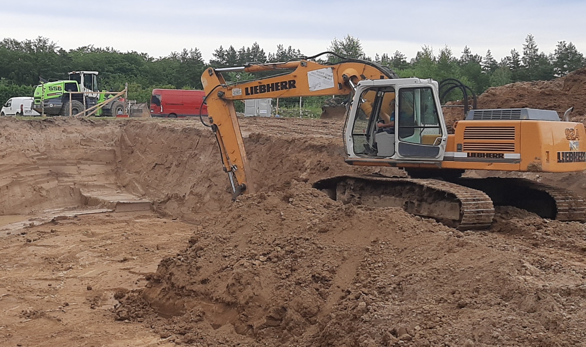Żółta koparka Liebherr 924 w trakcie prac ziemnych, widoczny wykop z betonowymi elementami oraz inne maszyny w tle, dzień pochmurny.
