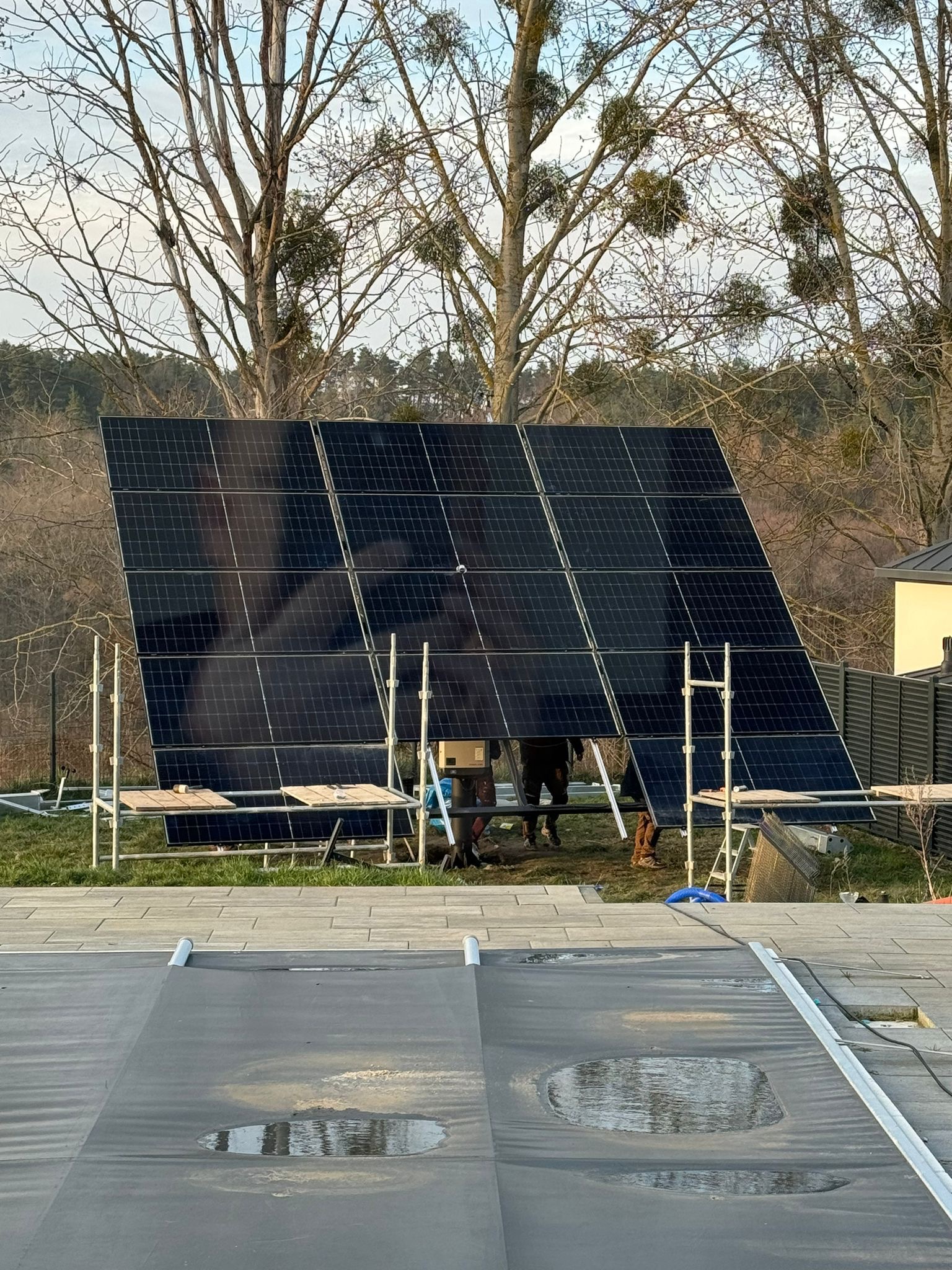 Montaż paneli słonecznych na stelażu w ogrodzie, widoczne rusztowania i ekipa montażowa, w tle drzewa bez liści i fragment pokrywy basenowej z kałużami.