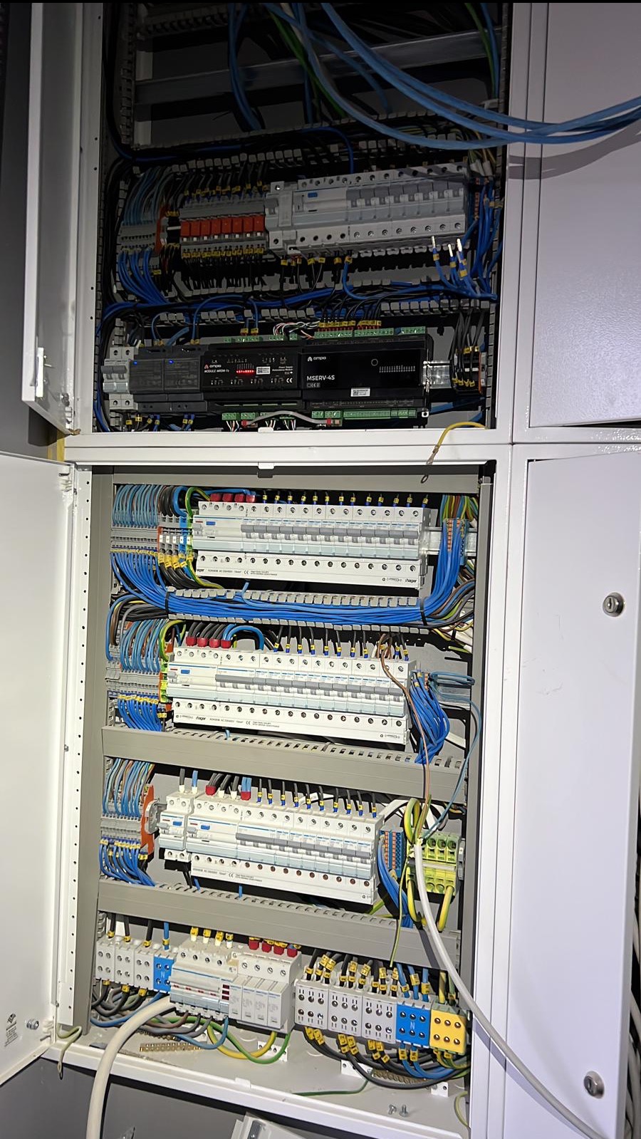 Otwarta szafa elektryczna z widocznymi bezpiecznikami, przewodami w kolorach niebieskim, żółtym i zielonym, oraz urządzeniem MSERV-4S, zainstalowana w pomieszczeniu.