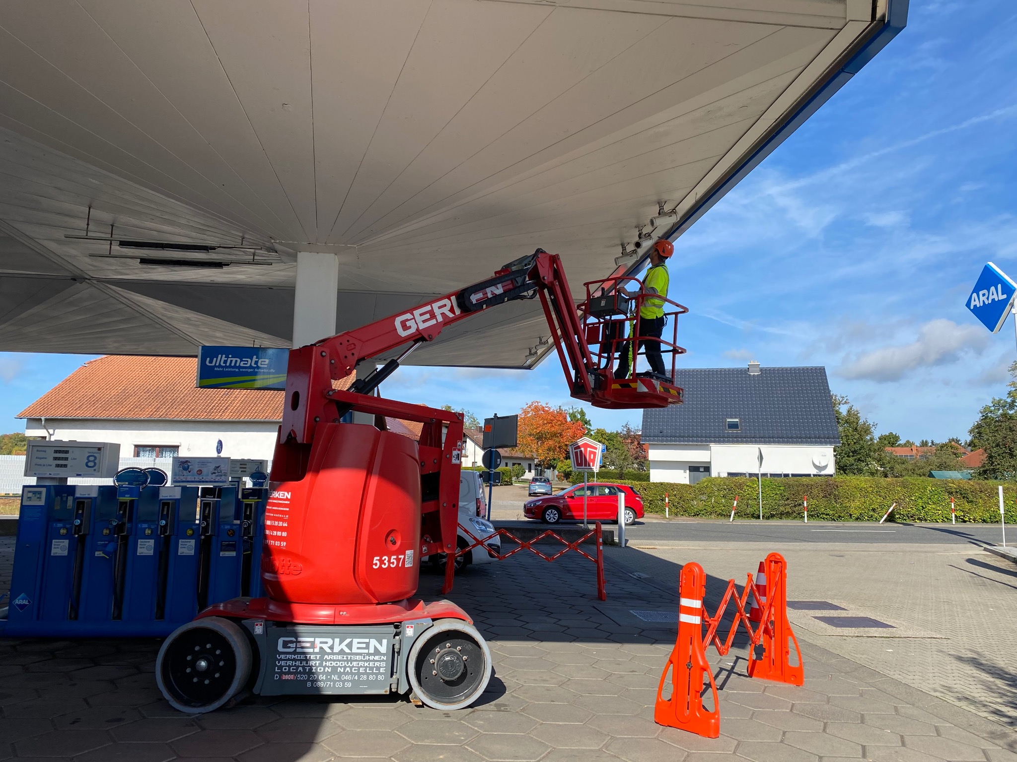 Pracownik w pomarańczowym kasku i żółtej kamizelce na czerwonym podnośniku koszowym Gerken, wykonujący prace konserwacyjne lub instalacyjne pod zadaszeniem stacji benzynowej Aral w słoneczny dzień.