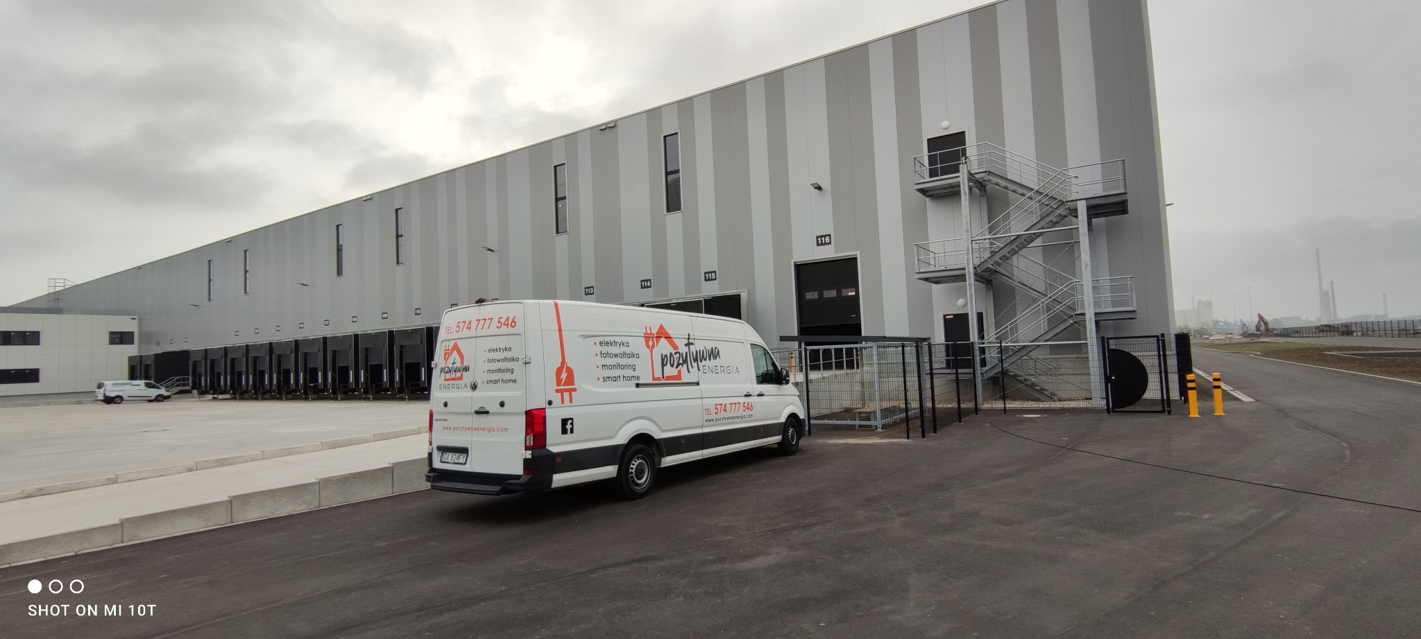 Biały van z logo firmy oferującej usługi elektryczne, fotowoltaiczne i inteligentnego domu zaparkowany na tle magazynu z rampami załadunkowymi i metalową klatką schodową.