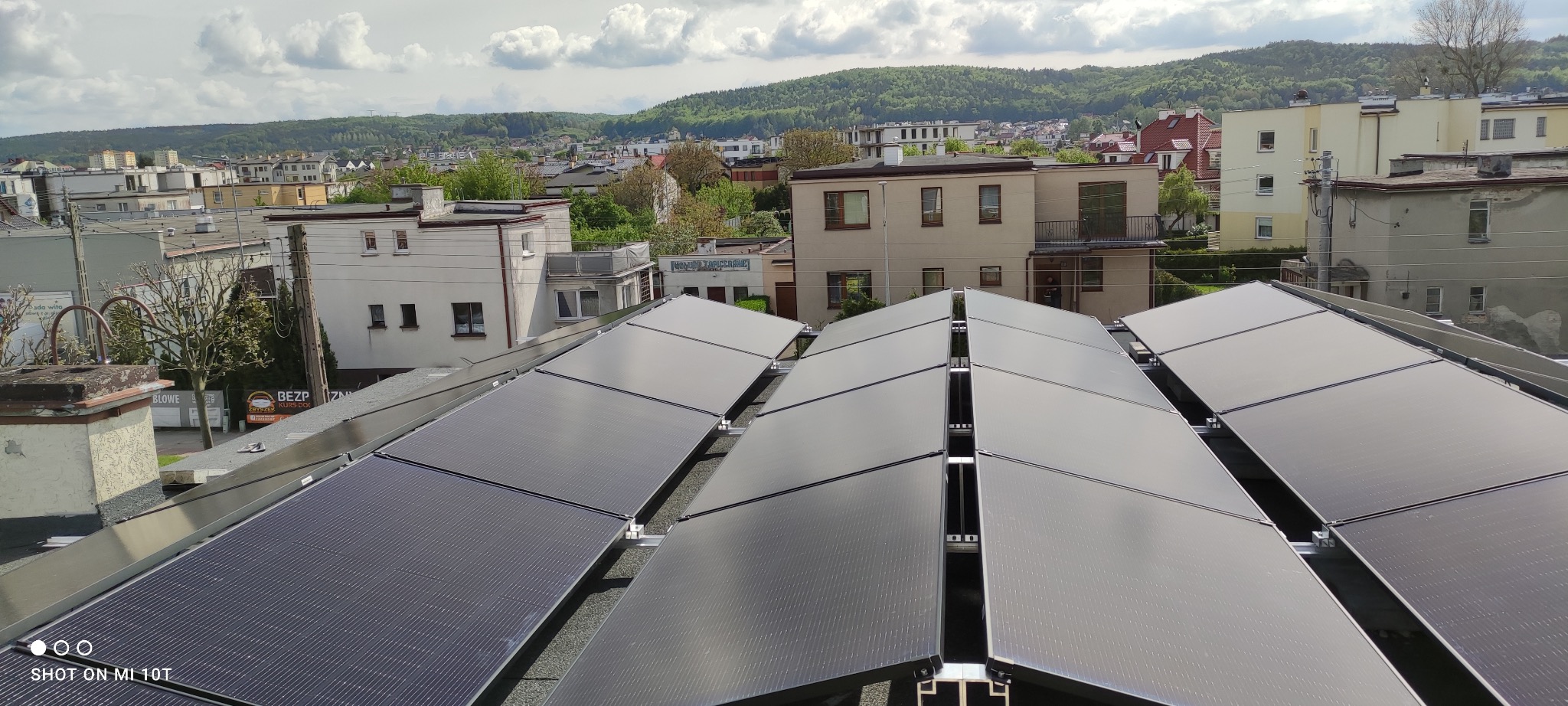 Widok z góry na panele słoneczne na dachu budynku w tle zabudowa miejska i zalesione wzgórza w pochmurny dzień.