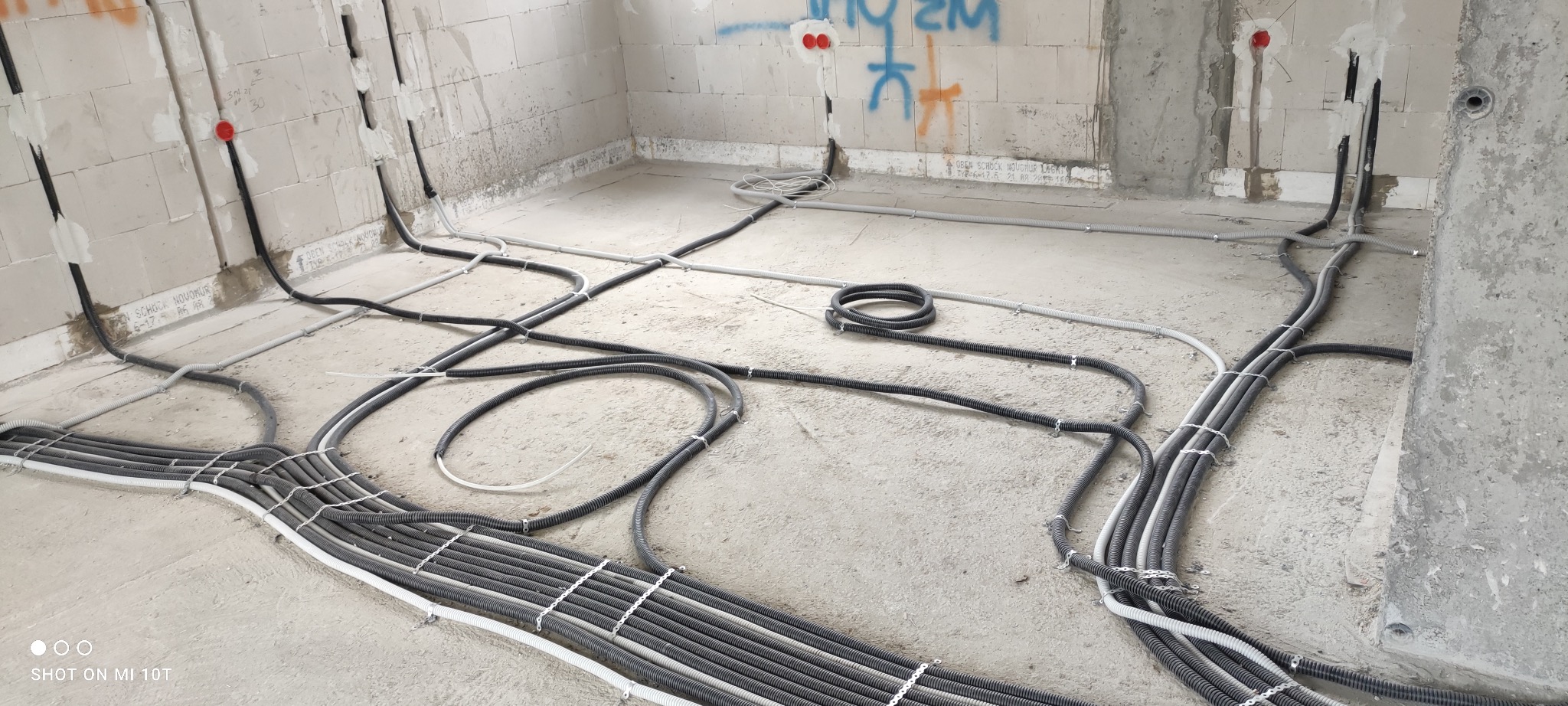 Wnętrze surowego budynku w trakcie prac instalacyjnych; widoczne liczne kable elektryczne w peszlach, prowadzone po podłodze i ścianach, mocowane uchwytami; ściany z betonu komórkowego z widocznymi...