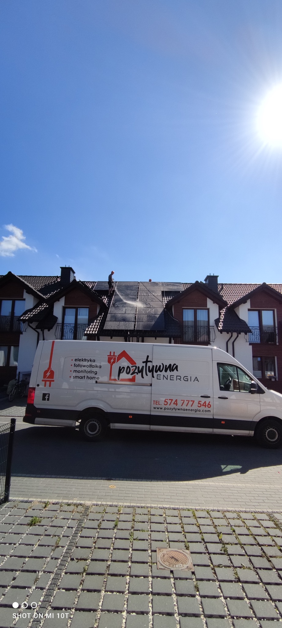 Czyszczenie paneli słonecznych na dachu dwupiętrowego domu, widoczna ekipa z myjką ciśnieniową, w tle błękitne niebo i firmowy bus z logo 'pozytywna energia' na ulicy.
