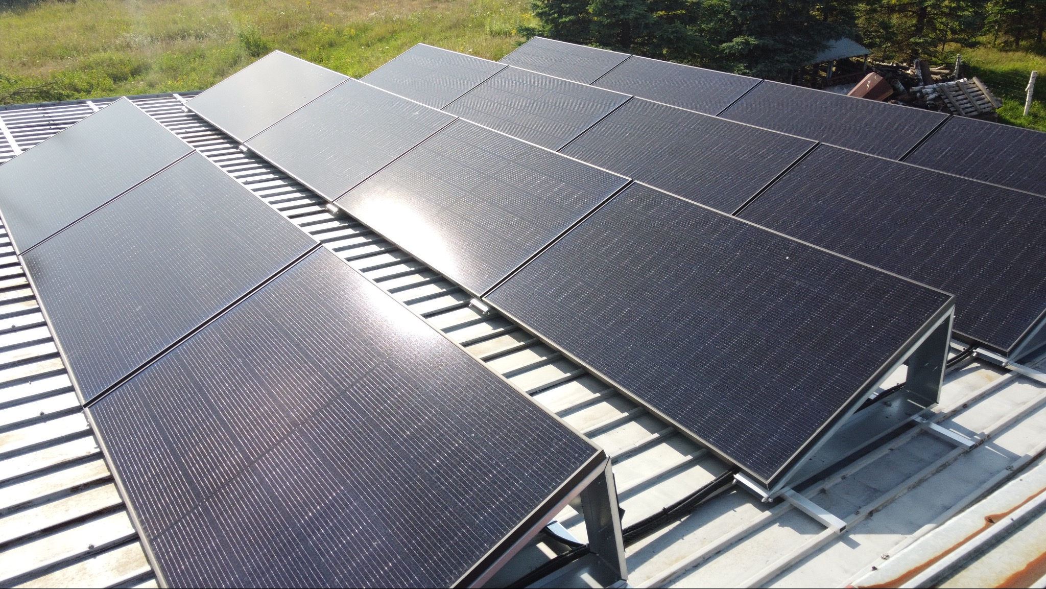 Rząd paneli słonecznych na dachu z blachy trapezowej, widoczne mocowania i kable, w tle zieleń drzew i trawy.