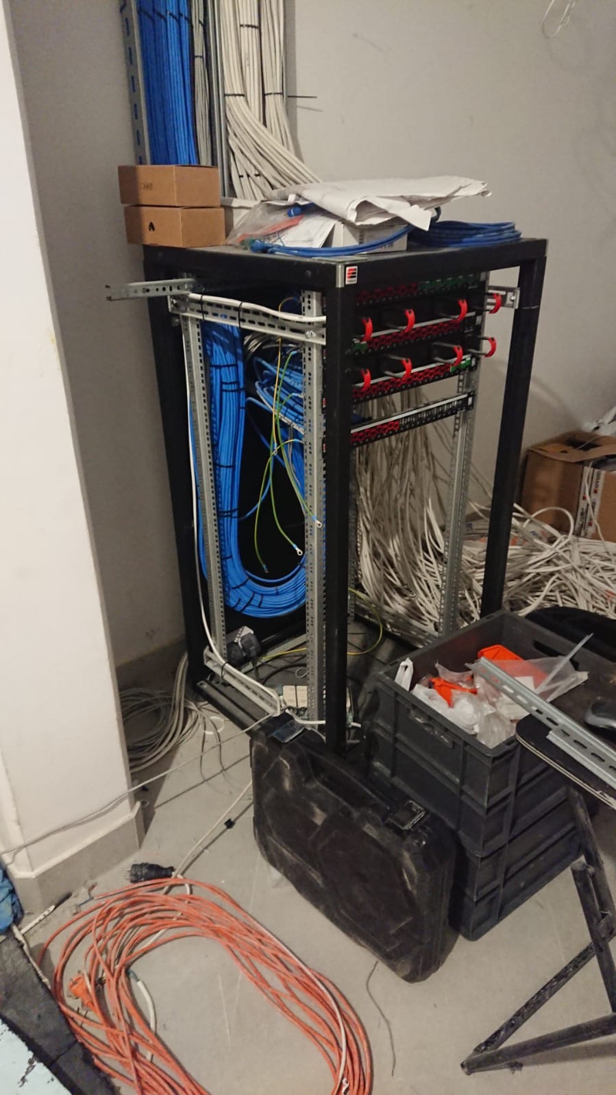 Stojak teleinformatyczny z ułożonymi kablami sieciowymi i elektrycznymi w trakcie instalacji, widoczne przewody, skrzynki i dokumentacja techniczna na górze, pomarańczowy przedłużacz na podłodze.