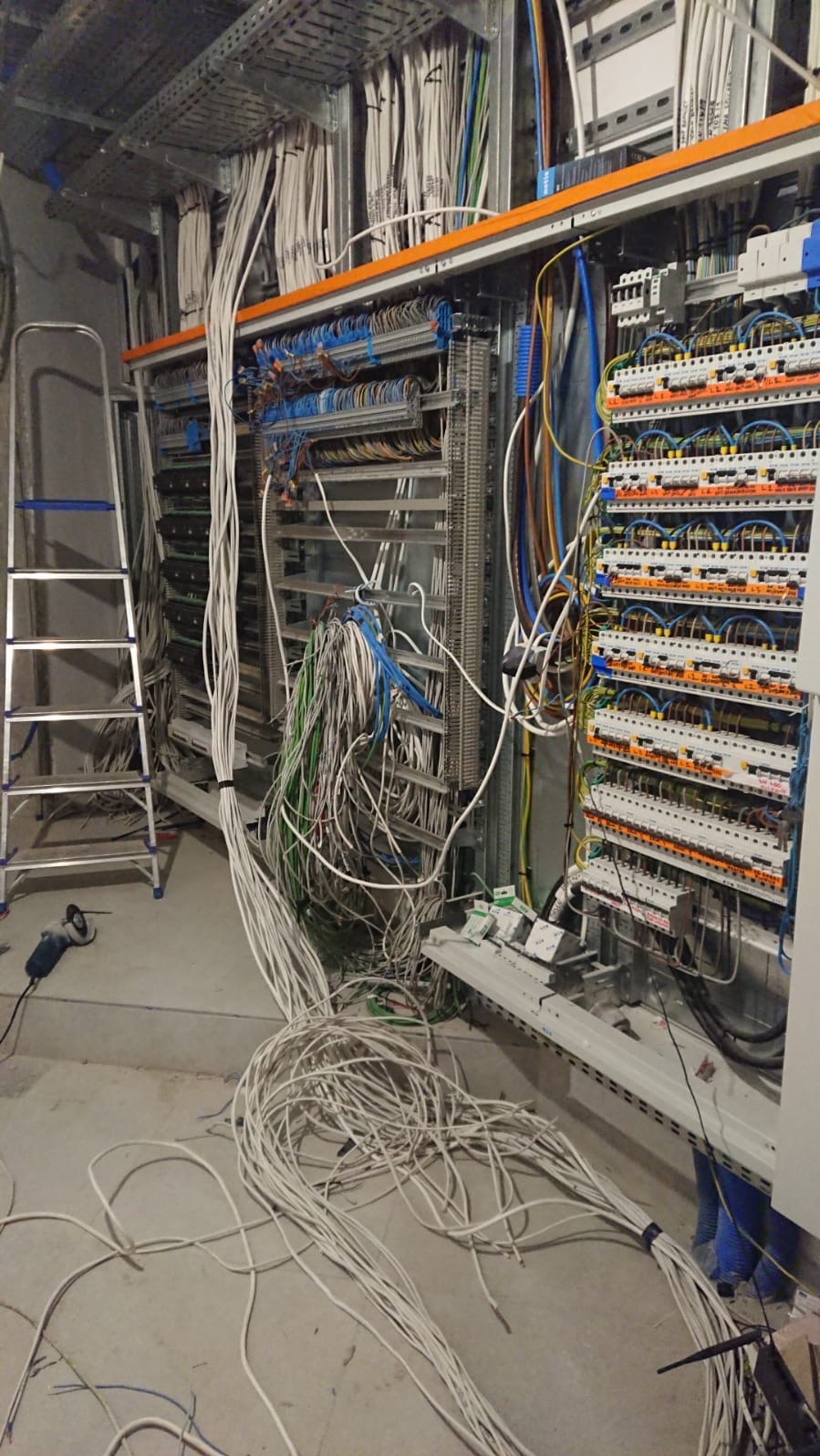 Rozbudowana instalacja elektryczna w trakcie montażu: liczne przewody, rozdzielnice, drabina i szlifierka kątowa na surowym betonowym podłożu.