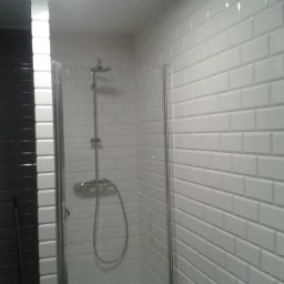 ZAKŁAD REMONTOWO-BUDOWLANY ANDRZEJ FORYŚ - Kabina prysznicowa z białymi, prostokątnymi płytkami ułożonymi w stylu cegiełki, kontrastująca z ciemną ścianą obok, widoczny zestaw prysznicowy.